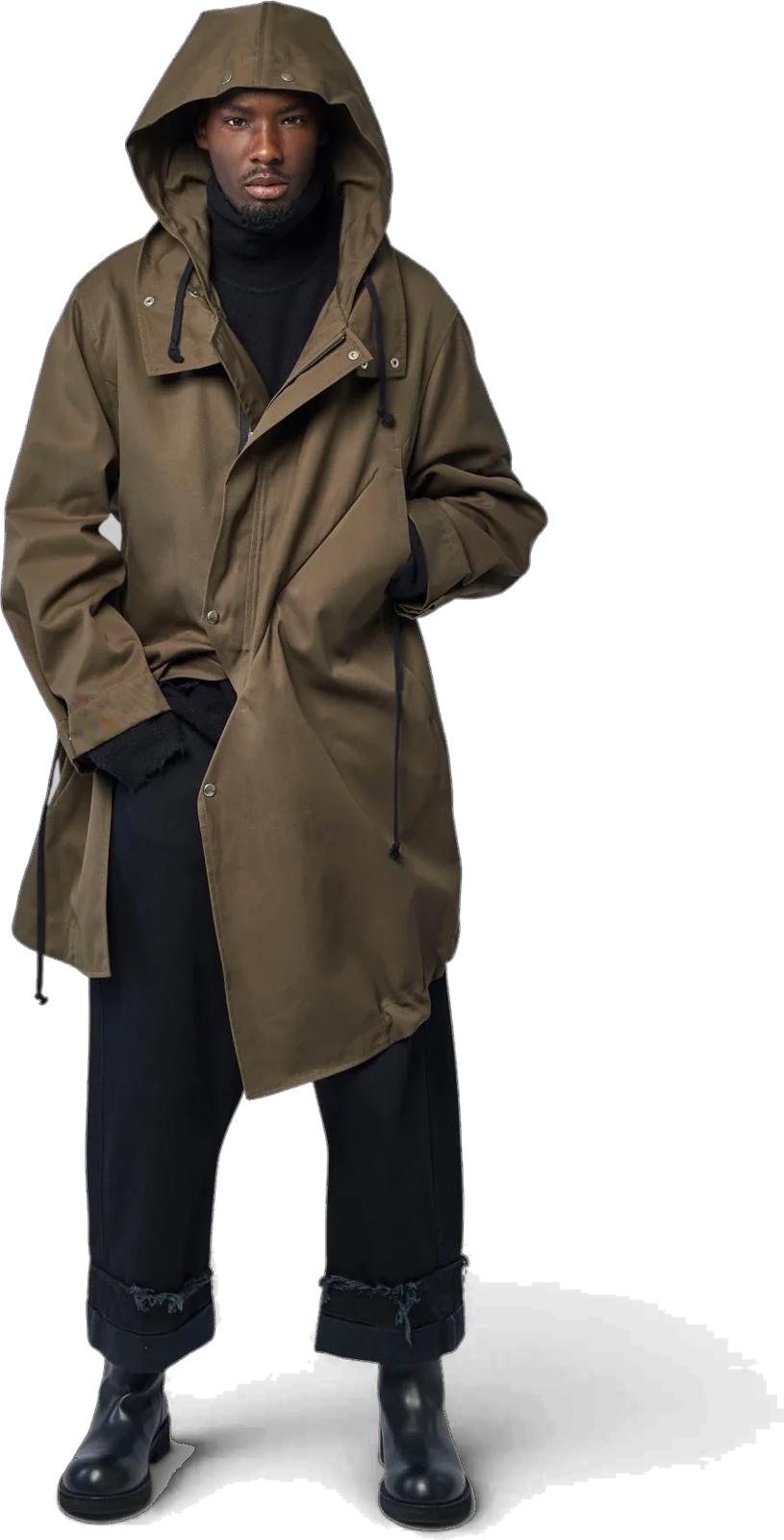 Parka 20 worn with the hood up: long olive cotton, drawcords at the collar, over black trousers turned up at the hem.